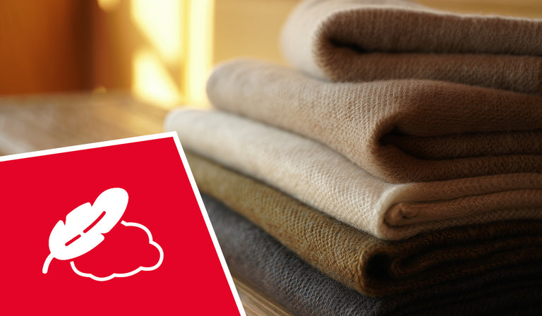Ein Stapel aus sechs weichen, gefalteten Stoffen in natürlichen Beige- und Brauntönen liegt auf einer Holzoberfläche im warmen Licht. Im Vordergrund links ist ein rotes Feld mit einem weißen Symbol eingeblendet – es zeigt eine Feder und eine Wolke, was für Weichheit oder Leichtigkeit steht.