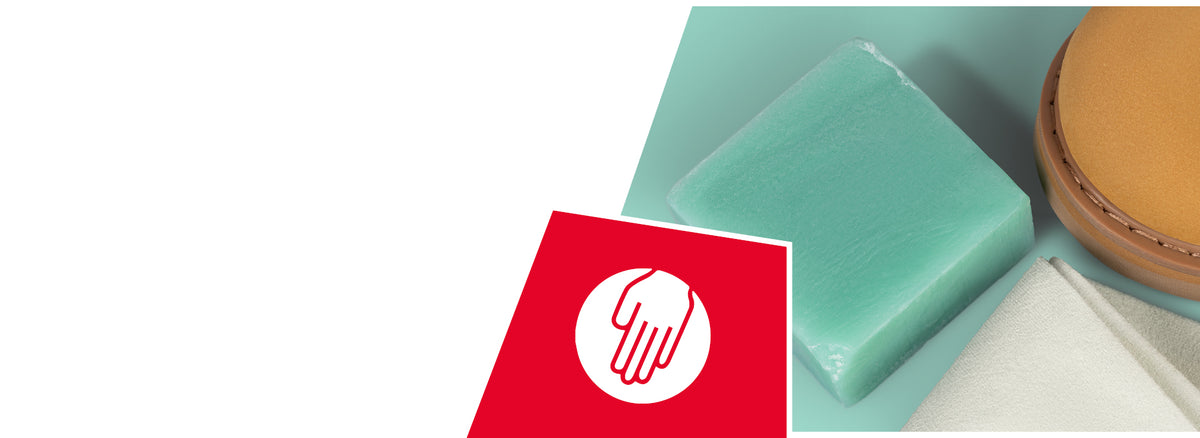 Detailaufnahme eines Pflege-Sets für Schuhe: Zu sehen sind ein grüner Reinigungsgummi, ein beigefarbenes Tuch und die Vorderkappe eines hellbraunen Schuhs. In der linken unteren Ecke befindet sich ein rotes Quadrat mit einem weißen Symbol – eine Hand auf einem Kreis –, das auf einfache manuelle Pflege hinweist.
