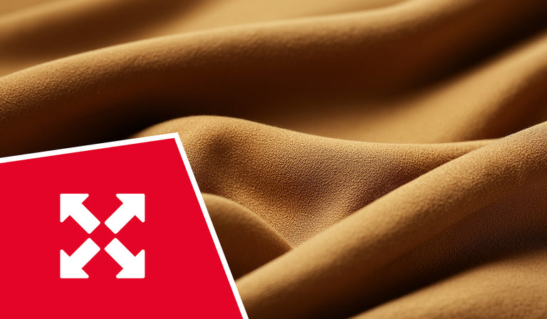 Nahaufnahme eines cognacbraunen, elastisch wirkenden Stoffes mit feiner Struktur, der in sanften Wellen liegt. In der unteren linken Ecke befindet sich ein rotes Quadrat mit einem weißen Symbol aus vier auseinander zeigenden Pfeilen – ein Hinweis auf Dehnbarkeit oder Flexibilität des Materials.