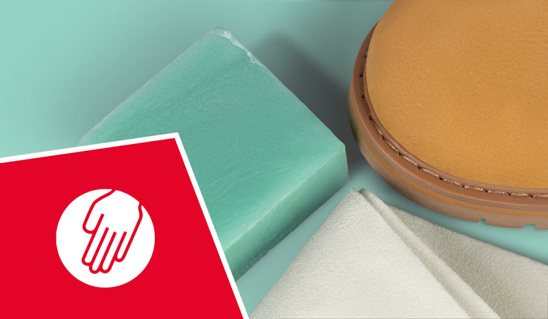 Detailaufnahme eines Pflege-Sets für Schuhe: Zu sehen sind ein grüner Reinigungsgummi, ein beigefarbenes Tuch und die Vorderkappe eines hellbraunen Schuhs. In der linken unteren Ecke befindet sich ein rotes Quadrat mit einem weißen Symbol – eine Hand auf einem Kreis –, das auf einfache manuelle Pflege hinweist.