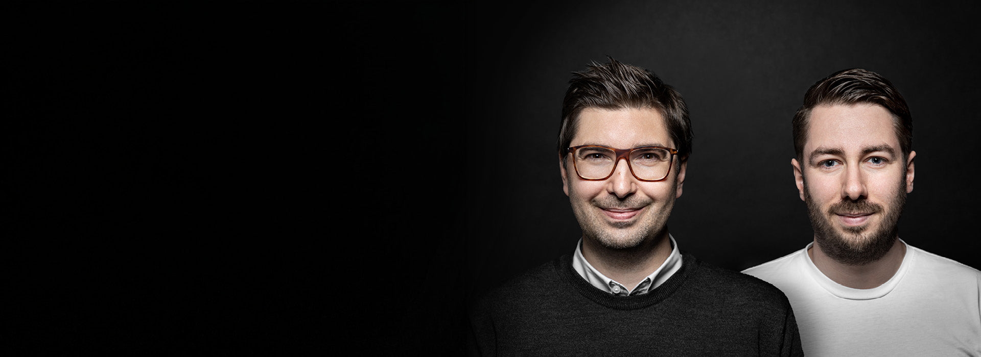Zwei Männer stehen vor dunklem Hintergrund nebeneinander. Der Mann links trägt eine Brille, ein weißes Hemd und einen dunklen Pullover. Der Mann rechts hat einen gepflegten Bart und trägt ein weißes T-Shirt. Beide blicken freundlich in die Kamera.