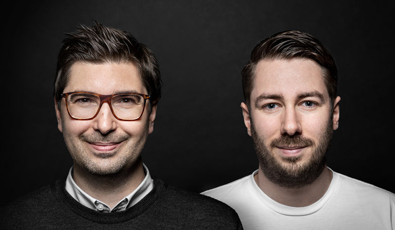 Zwei Männer stehen vor dunklem Hintergrund nebeneinander. Der Mann links trägt eine Brille, ein weißes Hemd und einen dunklen Pullover. Der Mann rechts hat einen gepflegten Bart und trägt ein weißes T-Shirt. Beide blicken freundlich in die Kamera.
