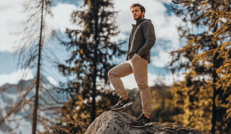 Ein junger Mann mit Bart steht selbstbewusst auf einem großen Felsblock in einer herbstlichen Waldlandschaft. Er trägt eine dunkelgraue Jacke und beige Hosen, dazu dunkle Sneaker. Im Hintergrund sind Nadelbäume und Laubbäume in warmen Herbstfarben zu sehen, der Himmel ist teilweise bewölkt. Die Szene vermittelt eine entspannte Outdoor-Atmosphäre bei goldenem Licht.