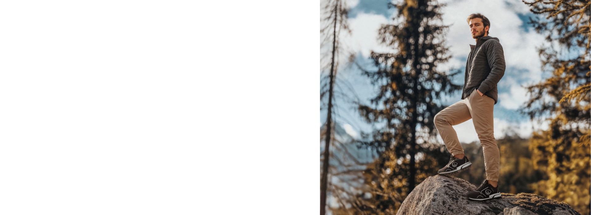 Ein junger Mann mit Bart steht selbstbewusst auf einem großen Felsblock in einer herbstlichen Waldlandschaft. Er trägt eine dunkelgraue Jacke und beige Hosen, dazu dunkle Sneaker. Im Hintergrund sind Nadelbäume und Laubbäume in warmen Herbstfarben zu sehen, der Himmel ist teilweise bewölkt. Die Szene vermittelt eine entspannte Outdoor-Atmosphäre bei goldenem Licht.