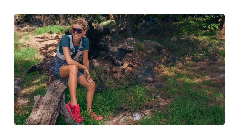 Lächelnde Frau mit Sonnenbrille in grauem T-Shirt und gemusterter Shorts mit roten Sneakers sitzt auf Baumstamm in natürlicher Waldumgebung mit Steinen und Gras.
