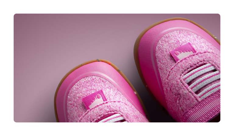 Nahaufnahme zweier pinker Giesswein Kinderschuhe mit Merino-Obermaterial, elastischen Schnürsenkeln und honigfarbener Sohle vor rosa Hintergrund.