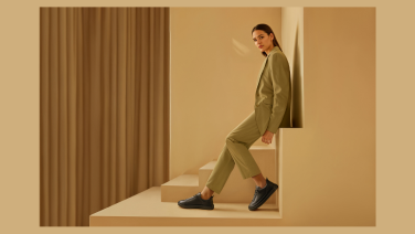 Junge Frau in olivgrünem Anzug sitzt auf einem beigen Podest in einem minimalistisch gestalteten Raum. Sie trägt schwarze Schuhe. Die Szene wirkt modern und elegant mit klaren geometrischen Formen.