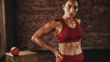 Sportliche Frau in roter Sportbekleidung steht selbstbewusst mit Hand an der Hüfte in einem Fitnessstudio vor Backsteinwand.