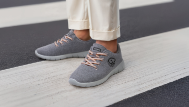 Füße in grauen Giesswein Sneakern mit orangefarbenen Schnürsenkeln stehen auf weißen Zebrastreifen. Die Person trägt weiße Hosen und die Schuhe zeigen das charakteristische Giesswein-Logo.