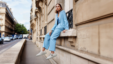 Frau in hellblauem Anzug sitzt entspannt auf einer Steinbalustrade vor klassischer Architektur. Sie trägt weiße Sneaker und lächelt in die Kamera. Im Hintergrund ist eine elegante Stadtstraße zu sehen.