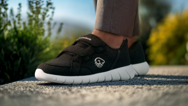 Nahaufnahme von Füßen in schwarzen Giesswein Sneakern mit weißer Sohle auf einem Kiesweg. Die Person trägt braune Hosen und im unscharfen Hintergrund sind grüne Pflanzen und Berge zu sehen.