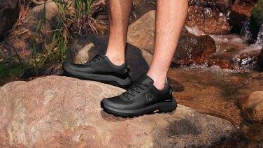 Zwei Füße in schwarzen Outdoor-Schuhen (Wool Trail X von Giesswein) stehen auf einem braunen Felsen am Ufer eines kleinen Bachs mit fließendem Wasser