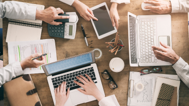 Mehrere Personen arbeiten gemeinsam an einem Tisch mit Laptops, Tablet, Taschenrechner und Unterlagen – eine geschäftige Teamatmosphäre.