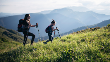 Zwei Wanderinnen mit Rucksäcken und Wanderstöcken steigen einen grasbewachsenen Berghang hinauf. Im Hintergrund erstreckt sich eine weite Berglandschaft mit mehreren Bergketten in bläulichen Farbtönen unter einem bewölkten Himmel.