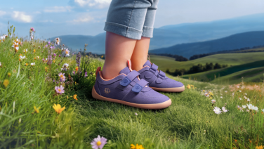 Lila Kinderschuhe mit Klettverschluss auf einer grünen Wiese mit bunten Wildblumen. Im Hintergrund erstreckt sich eine hügelige Landschaft mit sanften grünen Hügeln unter blauem Himmel.