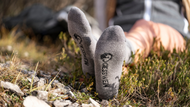 Person liegt auf dem Rücken im Gras, Füße in grauen Wandersocken mit schwarzem Logo und Schriftzug „GIESSWEIN“ nach vorn gestreckt. Im Hintergrund sind unscharf Wanderkleidung und Natur zu sehen.