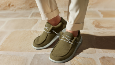 Nahaufnahme von Füßen in weißer Hose und olivgrünen Schuhen mit weißer Schnürung und heller Sohle. Die Person steht auf hellem Steinboden