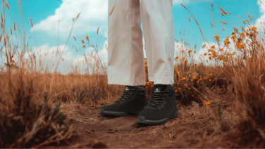 Person in weißer lockerer Hose und schwarzen Boots steht in einem trockenen Grasfeld unter blauem Himmel mit weißen Wolken