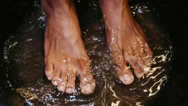 Barfüße einer Person, die in klarem, spritzenden Wasser steht. Wassertropfen und Spritzer sind in der Bewegung eingefroren, was die erfrischende und natürliche Atmosphäre betont.