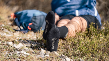 Eine Person liegt entspannt auf einem felsigen Untergrund in der Natur. Im Vordergrund sind die Füße mit schwarzen Giesswein Merino-Socken zu sehen.