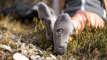Person liegt auf einer Wiese, im Vordergrund sind die Füße in grauen Socken mit Logo zu sehen.