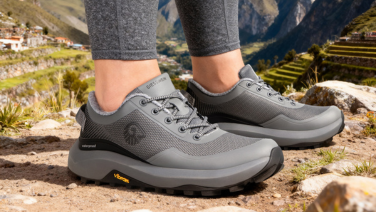 Nahaufnahme grauer Giesswein Outdoor-Schuhe mit wasserdichter Membran an den Füßen einer Frau; im Hintergrund eine felsige Berglandschaft mit terrassierten Feldern in Peru.