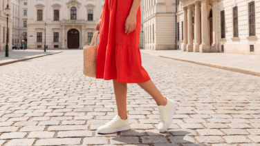 Frau in rotem Sommerkleid mit Korbtasche überquert eine Altstadtgasse aus Kopfsteinpflaster und trägt weiße Strick-Sneaker.