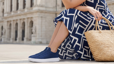 Frau in einem blau-weiß gemusterten Sommerkleid sitzt auf einer Stufe vor einem historischen Stadtgebäude, trägt marineblaue Strick-Sneaker und hält einen geflochtenen Korbtaschen-Henkel in der Hand.