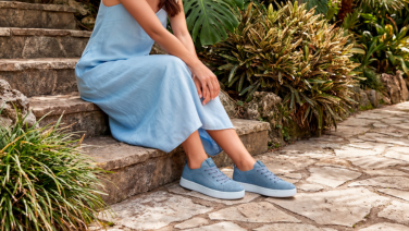 Frau in hellblauem Leinenkleid sitzt auf Steinstufen in einem mediterranen Garten und trägt hellblaue Strick-Sneaker.
