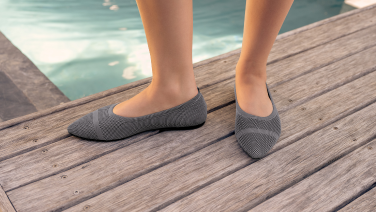 Füße in grauen Strick-Ballerinas auf einem hölzernen Steg neben einem Pool.