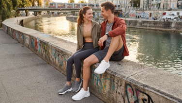 Paar sitzt lachend auf einer Mauer an einem Fluss in der Stadt, beide tragen graue Giesswein Merino Runner Sneaker