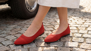 Nahaufnahme von Füßen in roten Giesswein Strick-Ballerinas, stehend auf einem Kopfsteinpflaster neben einem geparkten Auto.