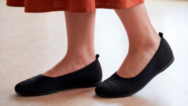 Nahaufnahme von Füßen in schwarzen Giesswein Strick-Ballerinas mit Zuglasche an der Fersenpartie, unter einem terrakottafarbenen Rock, auf hellem Boden.