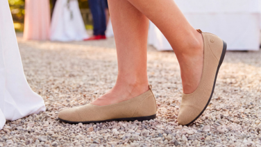 Nahaufnahme von Füßen in beigen Giesswein Strick-Ballerinas auf einem Kieselweg, im Hintergrund eine festlich gedeckte Tafel bei einer Feier.
