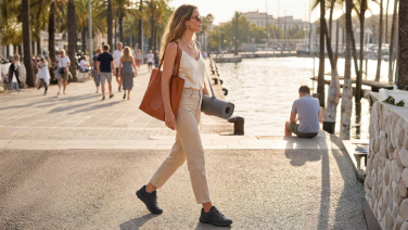 Frau mit Tasche und Yogamatte geht an einer Strandpromenade mit Palmen entlang, trägt schwarze Giesswein Wooltastic Sneaker.