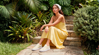Frau sitzt auf einer Steintreppe in einem üppigen Garten. Sie trägt ein gelbes Sommerkleid, ein weißes Haarband und cremefarbene Giesswein Sneaker.