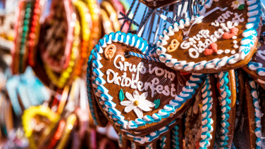 Mehrere bunt verzierte Lebkuchenherzen hängen dicht nebeneinander an einem Marktstand. Im Vordergrund ist ein Herz mit weißer Zuckerschrift und einer Blüte zu erkennen, umrandet von blau-weißem Zuckerguss.