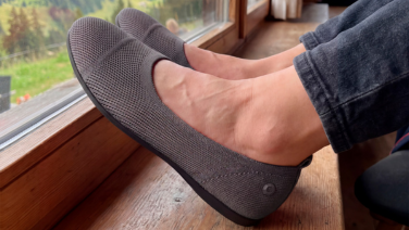 Nahaufnahme der Füße einer Person, die mit hochgekrempelter dunkler Hose auf einer Holzfensterbank abgelegt sind. Sie trägt taupefarbene, strukturierte Strick-Ballerinas mit dunkler Sohle. Im Hintergrund ist eine grüne Almwiese zu sehen.