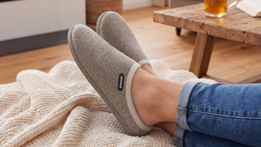 Nahaufnahme der übereinandergelegten Füße einer Person in Jeans, die auf einer cremefarbenen Strickdecke liegen, im Hintergrund ein Holztisch mit Getränk und Buch. Sie trägt graue Merino Wolle Pantoffeln mit hellem Rand.