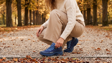 Eine Person in cremefarbenem Strickpullover und beiger Hose hockt auf einem Kiesweg mit Herbstlaub, im Hintergrund eine Allee. Sie trägt blaue Outdoor-Schuhe mit Drehverschluss und profilierter Sohle.