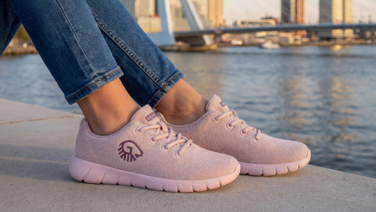 Nahaufnahme von zwei Füßen in rosafarbenen Wooltastic Sneakern von Giesswein, die auf einer Mauer vor einem Fluss mit Brücke im Hintergrund stehen.