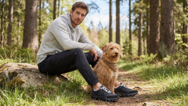 Ein Mann in grauem Hoodie und dunkler Hose sitzt auf einem Baumstamm im Wald neben einem Goldendoodle und trägt schwarze Giesswein Outdoor-Sneaker.