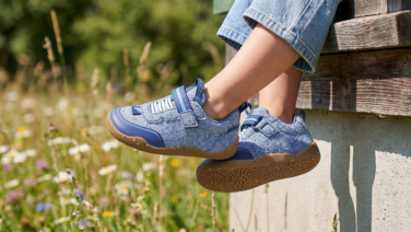 Nahaufnahme hellblauer Giesswein Kinder-Sneaker mit Klettverschluss und honigfarbener Sohle, getragen zu Jeans vor einer blühenden Sommerwiese.
