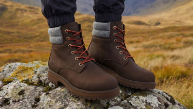 Nahaufnahme der Füße einer Person, die auf einem Felsen in einer herbstlichen Berglandschaft steht. Sie trägt braune Lederboots mit roten Schnürsenkeln, grauem Wollfilz-Kragen und profilierter Sohle.
