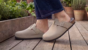 Nahaufnahme der Füße einer Person in dunkelblauer Jeans, die auf einer Holzterrasse steht. Sie trägt beigefarbene, Merino Clogs mit Schnallenriemen und Korksohle. Im Hintergrund stehen Kräutertöpfe und ein Hochbeet.