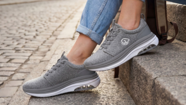 Nahaufnahme der Füße einer Person in umgekrempelter Jeans, die auf einer Steinstufe sitzt, daneben eine braune Ledertasche. Sie trägt graue, melierte Merino Wolle Sneaker mit weißer Sohle und Air-Cushion-Element in der Ferse.