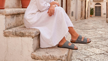 Frau in weißem Kleid sitzt auf einer Steintreppe in einer mediterranen Gasse und trägt graue Giesswein Sandalen mit zwei Riemen und Korkfußbett.