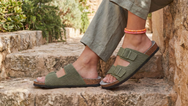 Füße in olivgrünen Strick-Sandalen mit zwei Schnallen und Korkfußbett, auf mediterranen Steinstufen, dazu eine olivgrüne Leinenhose und farbige Fußkettchen.