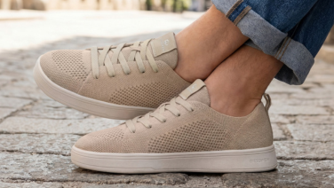 Nahaufnahme eines sandfarbenen Giesswein Sneakers mit strukturiertem Strickobermaterial und weißer Sohle. Die Person trägt helle Jeans und sitzt auf einem Kopfsteinpflaster.
