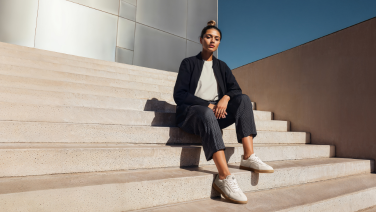 Eine Frau in dunkelblauem Blazer und grauer Hose sitzt auf Steinstufen vor moderner Architektur und trägt weiße Giesswein Merino-Sneaker.